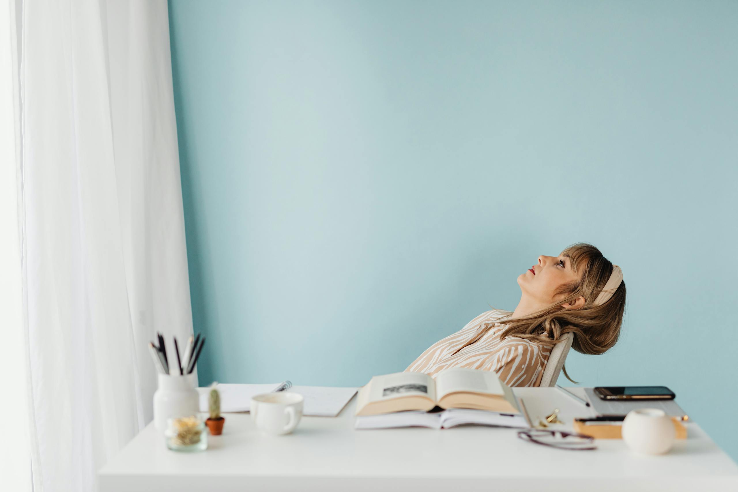 A woman is reclining at a desk with books, looking thoughtful and relaxed.