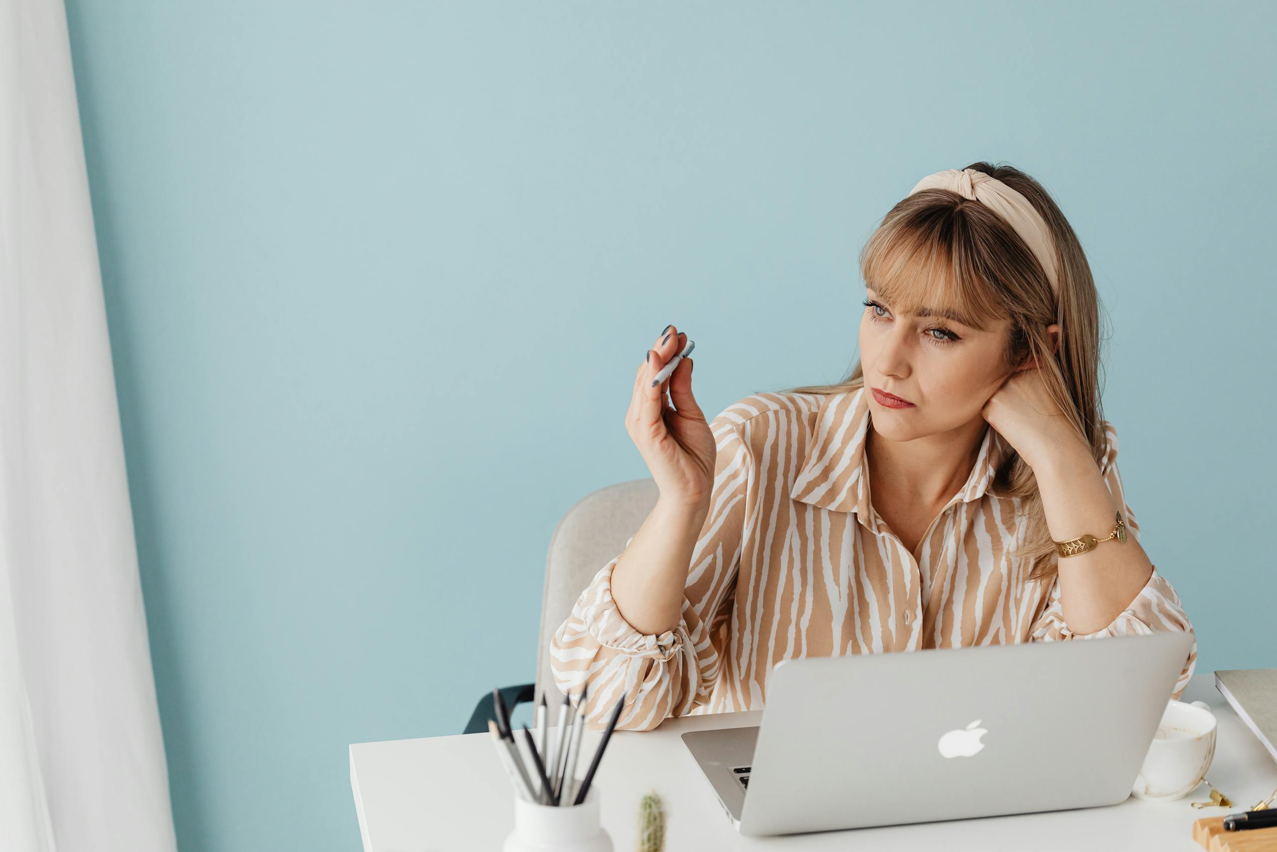 A woman relaxing in a home office setting, sitting with a laptop, fidgeting with a pencil, and enjoying a cup of coffee.